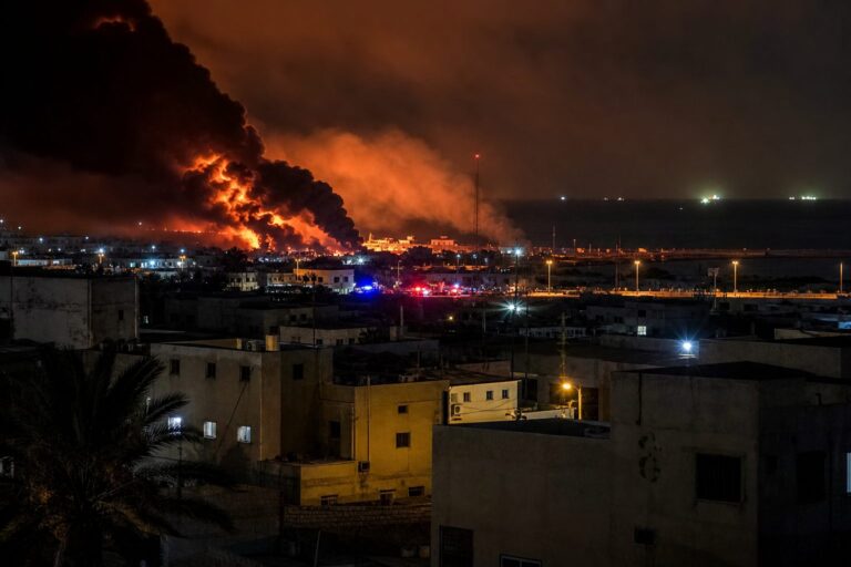 Large fire and black smoke rising behind buildings of a town at night
