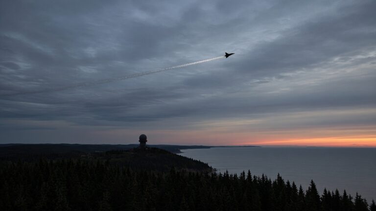 Fighter jet leaving a contrail over a dark forested coast with a radar dome at dusk