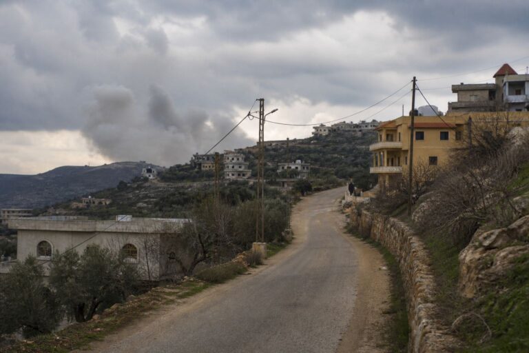 Smoke rising behind a hillside village road lined with power lines