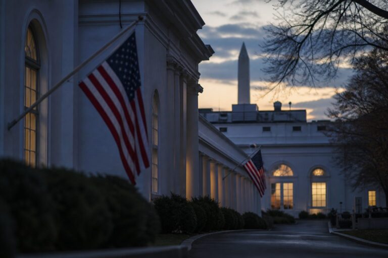 White House portico with U.S. flags and the Washington Monument at dusk