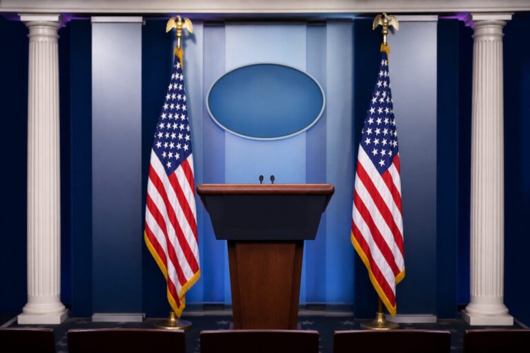 Empty White House-style briefing podium flanked by two U.S. flags