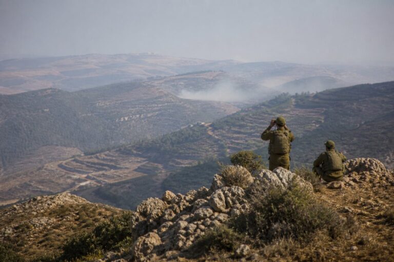 Two soldiers watching from a rocky ridge over terraced, hazy hills