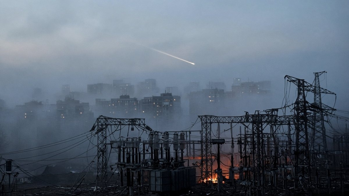Smoke rising over a dark city skyline at dawn above a damaged electrical substation with power lines