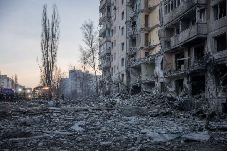 Rubble in front of a partly collapsed apartment block with emergency vehicles in the distance