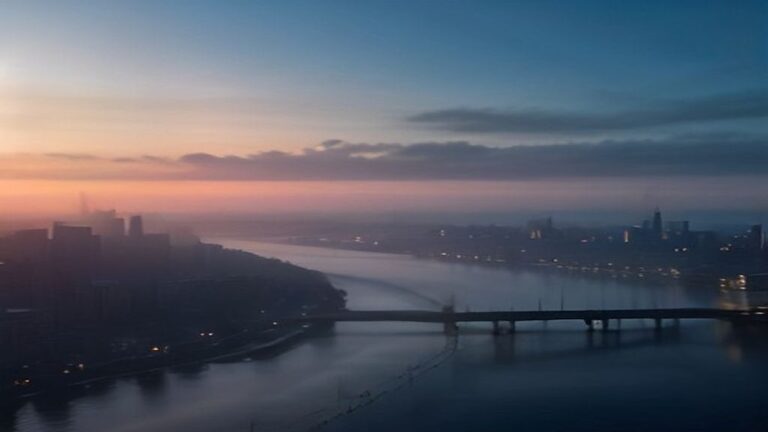 Dawn over a wide river city with a long bridge deck and thin smoke above apartment towers