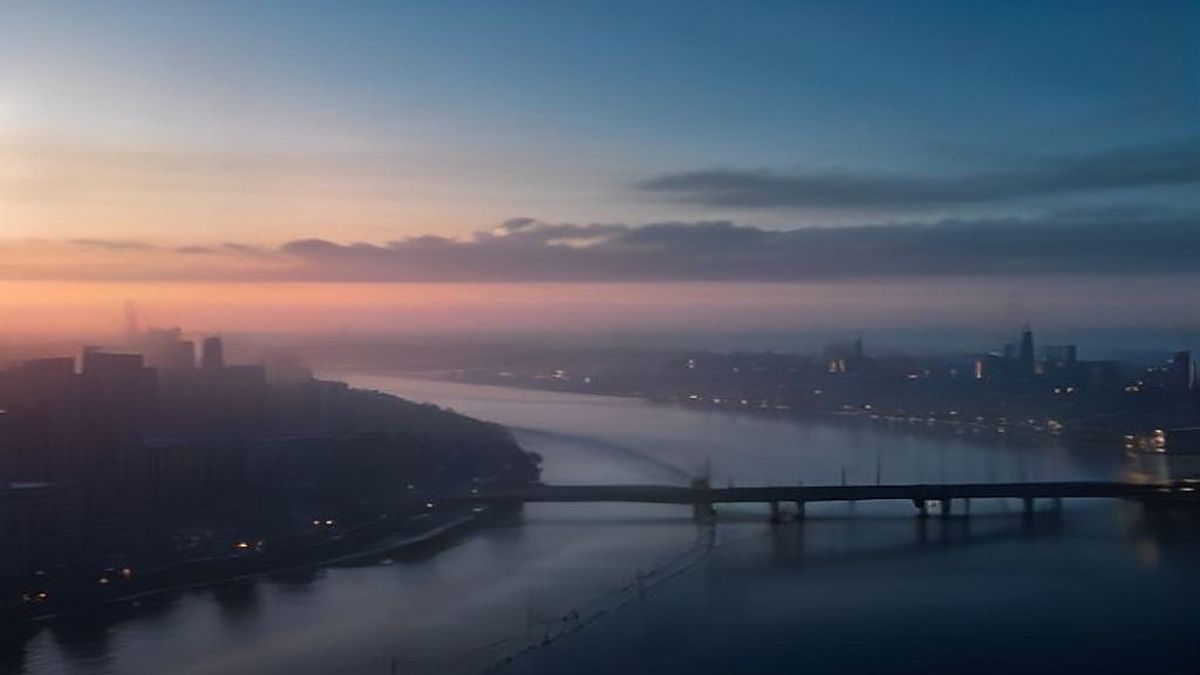 Dawn over a wide river city with a long bridge deck and thin smoke above apartment towers